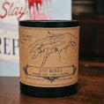 Load image into Gallery viewer, A black soy wax candle with a brown kraft paper label reading 'Just Buried' sits on a wooden table in front of a blurred book titled 'Kiss Slay' by Rachel Harrison.
