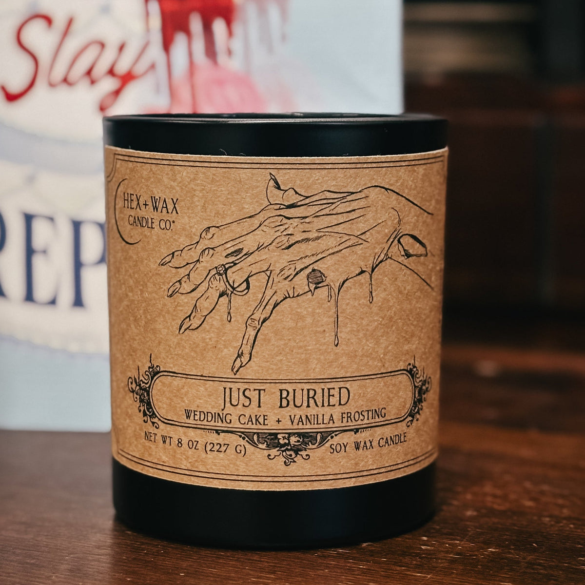 A black soy wax candle with a brown kraft paper label reading 'Just Buried' sits on a wooden table in front of a blurred book titled 'Kiss Slay' by Rachel Harrison.
