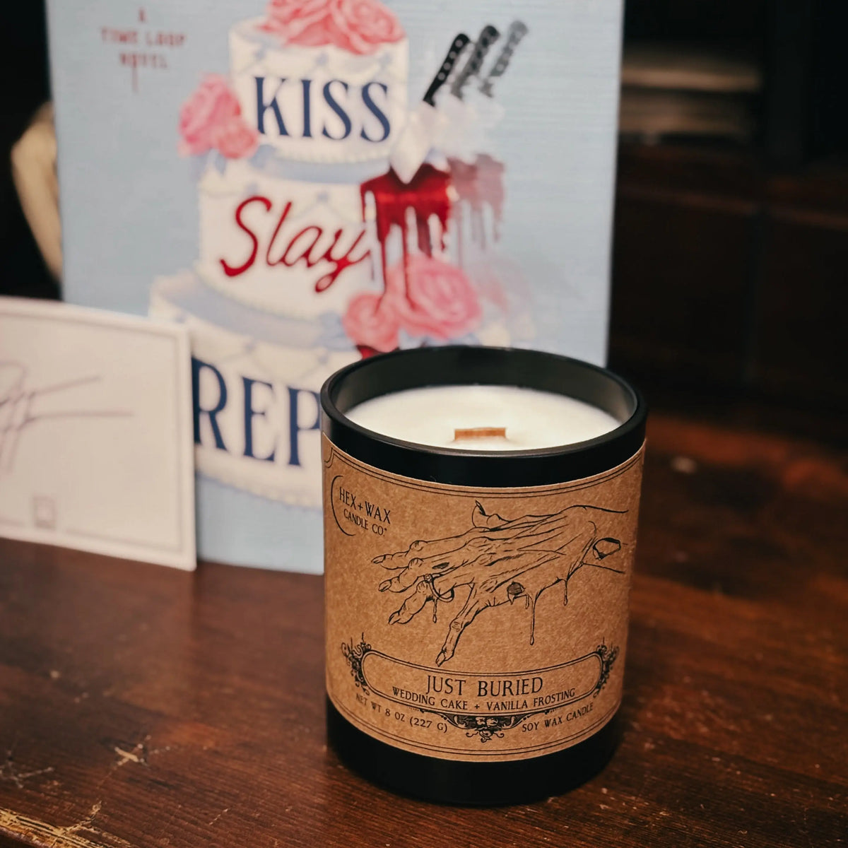 A black candle jar with a kraft paper label reading 'Just Buried' sits on a wooden table in front of a book cover for Rachel Harrison's 'Kiss Slay Rep' novel.